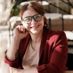 Person with glasses and a red jacket smiling at a table with a drink.