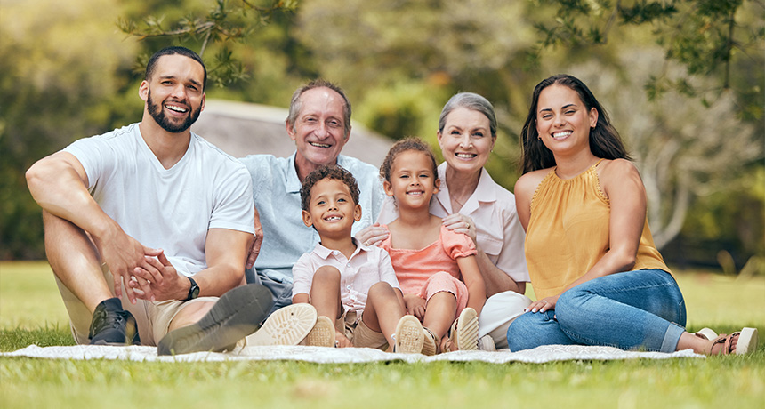 A family of six sits together on a picnic blanket in a park, smiling at the camera. The group includes adults, children, and an elderly person.