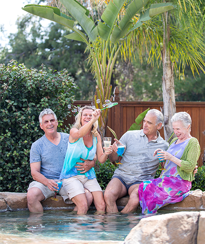 Four older adults sit by a pool, smiling and holding drinks. A woman in a blue top blows bubbles. Palm trees and greenery are in the background.
