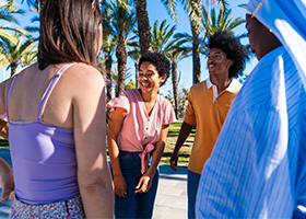 A group of four people stands outdoors, smiling and talking, with palm trees in the background on a sunny day.