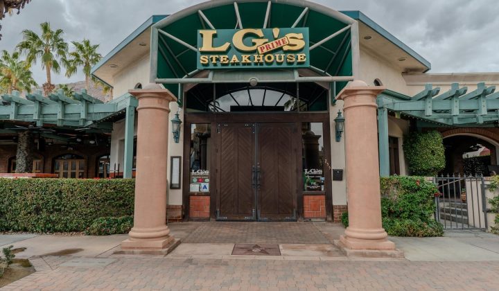 Entrance of LG's Prime Steakhouse with a green awning and wooden double doors, flanked by columns and greenery under a cloudy sky.