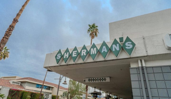 Sign reading "SHERMAN'S" on a building facade, with diamond-shaped tiles and palm trees in the background.