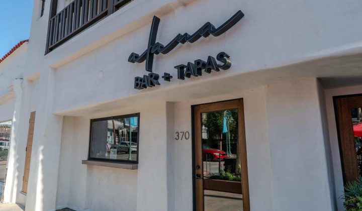 White building facade with a sign reading "Alana Bar + Tapas" and the address number 370. A brown door and window are visible, with a reflection of the street in the glass.