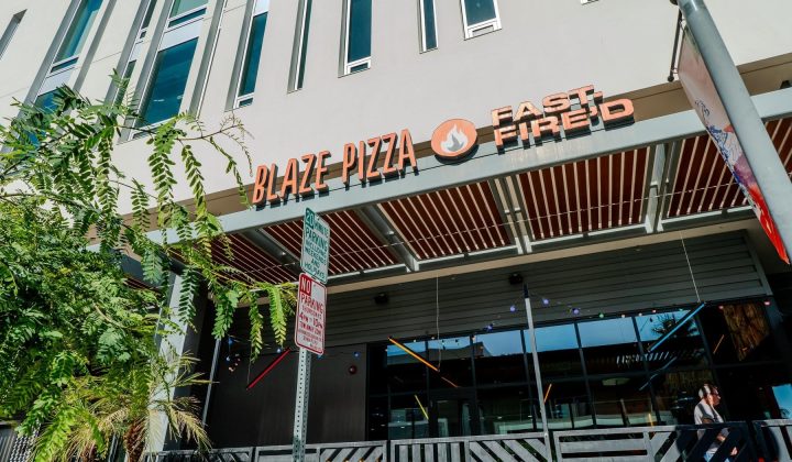 Street-level view of a Blaze Pizza restaurant with "Fast Fired" signage. A tree and a street sign are visible in the foreground.