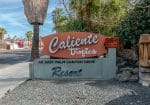 Sign for Caliente Tropics Resort at 411 East Palm Canyon Drive, surrounded by palm trees and a tiki statue.