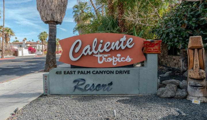 Sign for Caliente Tropics Resort at 411 East Palm Canyon Drive, surrounded by palm trees and a tiki statue.