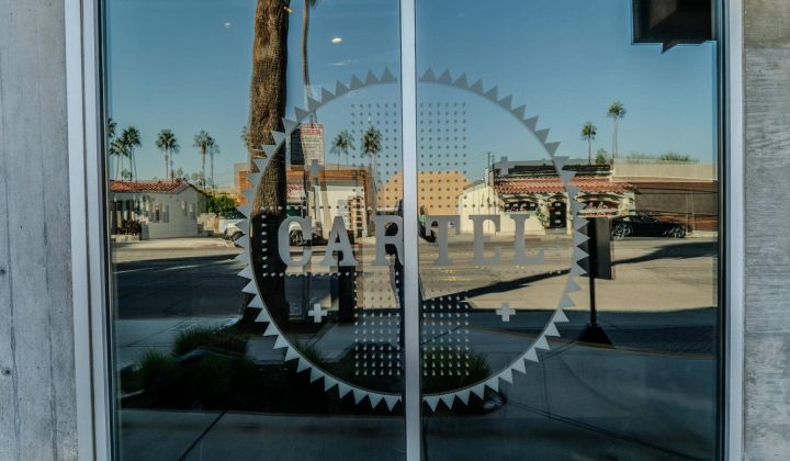 Glass door with "CARTEL" logo, reflecting palm trees, street signs, and buildings in the background.