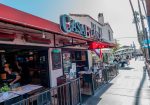 Restaurant with a red canopy and "Casa Blanca" sign, open for business. Outdoor tables and pedestrians on a sunny street with palm trees in the background.
