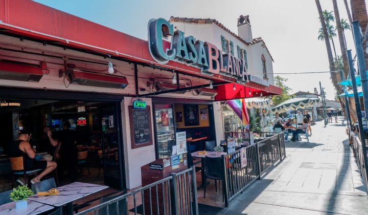 Restaurant with a red canopy and "Casa Blanca" sign, open for business. Outdoor tables and pedestrians on a sunny street with palm trees in the background.