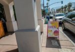 A sidewalk with a sign reading "Cheeky’s OPEN" next to a row of parked cars. Background shows palm trees and a traffic light.