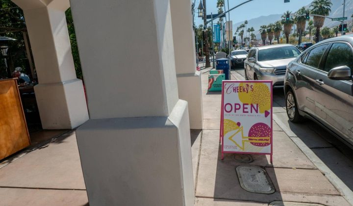 A sidewalk with a sign reading "Cheeky’s OPEN" next to a row of parked cars. Background shows palm trees and a traffic light.