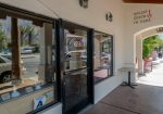 Entrance of Cheeky's restaurant with a glass door and a "Hello! Check in here" sign on the white wall. Storefront windows display items. Sidewalk and street view with palm trees outside.