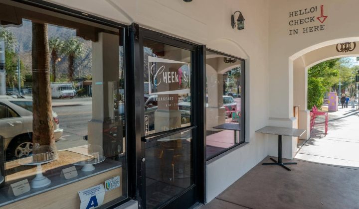 Entrance of Cheeky's restaurant with a glass door and a "Hello! Check in here" sign on the white wall. Storefront windows display items. Sidewalk and street view with palm trees outside.