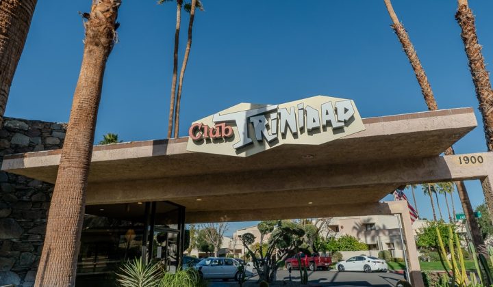 Entrance of Club Trinidad, featuring a retro-style sign and surrounded by palm trees. Cars are parked nearby.