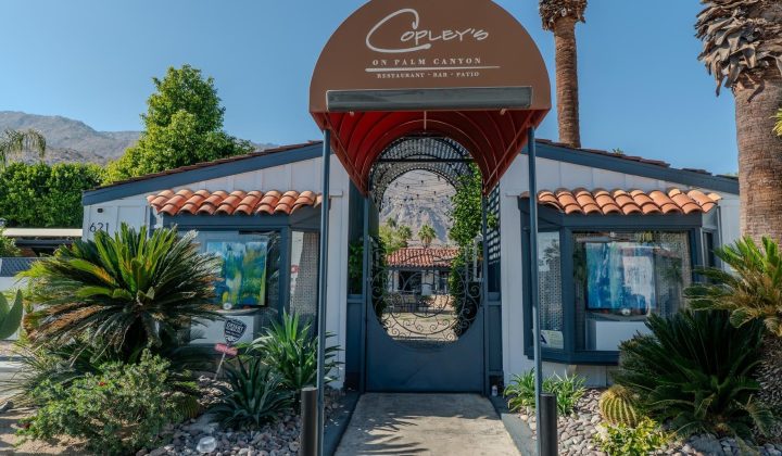 Entrance of Copley's on Palm Canyon, a restaurant with a gated archway, red and white building, surrounded by desert landscaping and palm trees.