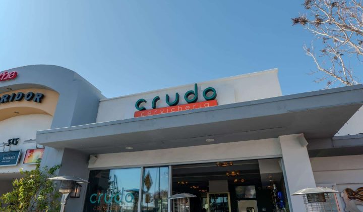 Facade of a restaurant named "Crudo Cevicheria" with a modern design and outdoor heaters. The sky is clear and there are trees nearby.