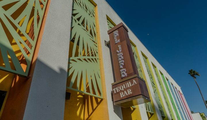 Building exterior with a colorful facade featuring palm leaf patterns. A vertical sign reads "El Jefe Tequila Bar." The sky is clear and there's a single palm tree in the background.