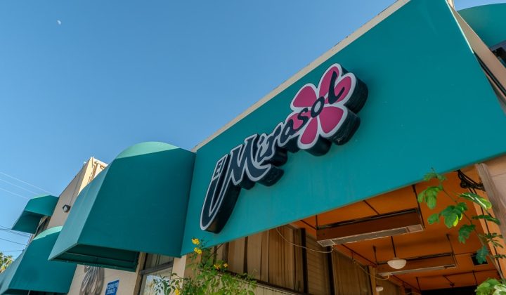 Exterior of a building with a teal awning and a sign featuring the word "Mirasol" and a flower design. Sky is clear with a small visible moon.