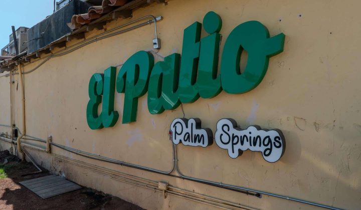Green and white "El Patio Palm Springs" sign on a beige wall with visible wiring and pipes.