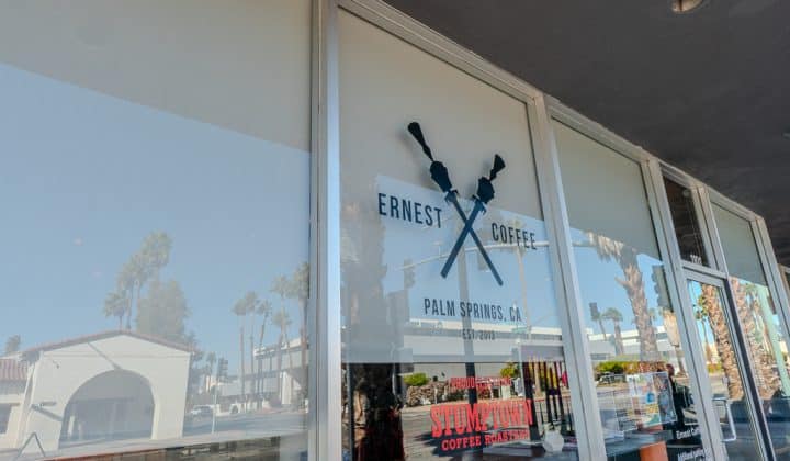 Glass storefront with palm trees reflected. "Ernest Coffee" sign featuring two crossed rifles, Palm Springs, CA.