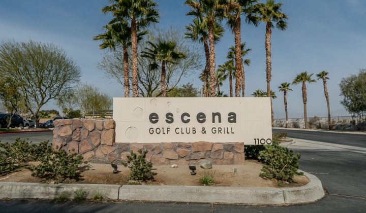 Sign for Escena Golf Club & Grill surrounded by palm trees and desert landscaping.