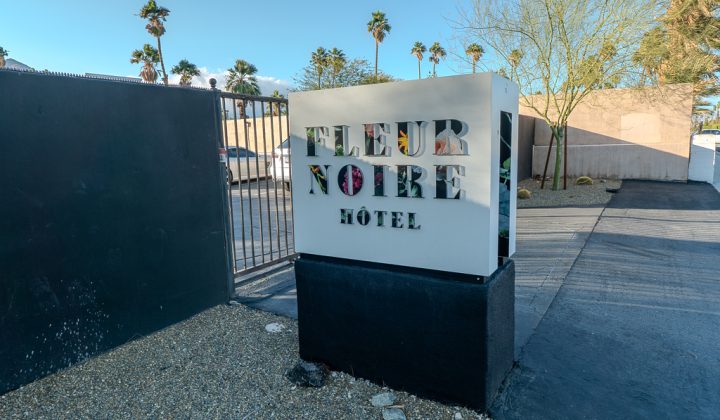 Sign reads "Fleur Noire Hôtel" in front of a gated entrance. The surrounding area features desert landscaping with gravel and palm trees.