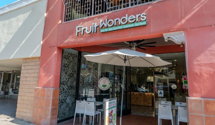 Exterior of a juice bar called "Fruit Wonders" with an orange facade. White chairs and tables are set under a large umbrella outside the entrance.
