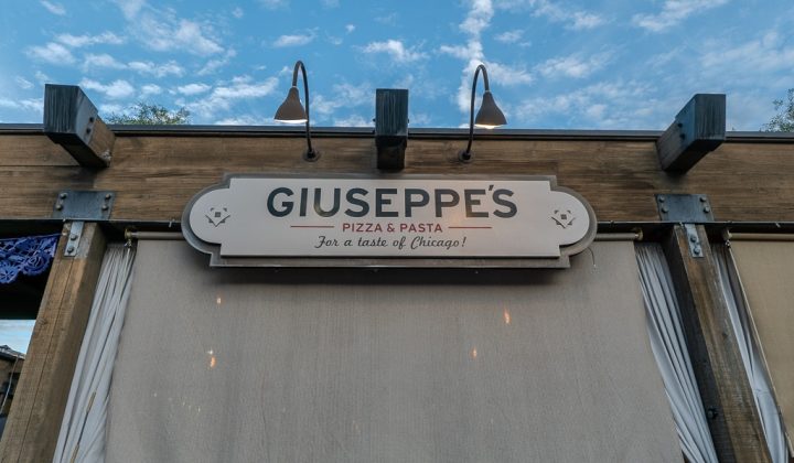 Sign reading "Giuseppe's Pizza & Pasta, For a taste of Chicago!" mounted on a wooden structure with lights above, against a partly cloudy sky.