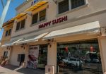 Street view of a sushi restaurant named "Happy Sushi" with large windows, yellow awnings, and a display poster featuring sushi dishes. Several parked cars and a potted plant are visible.