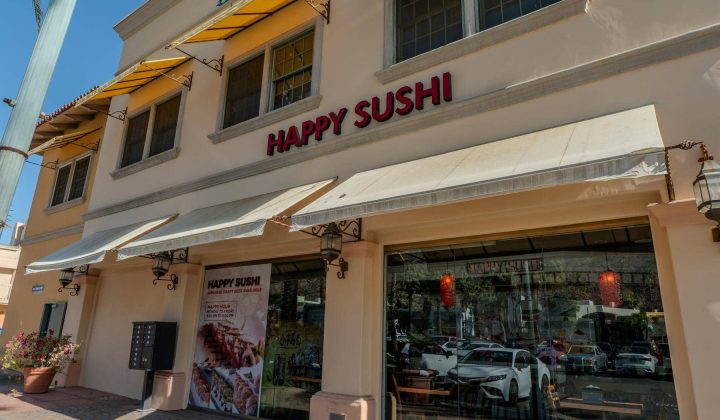 Street view of a sushi restaurant named "Happy Sushi" with large windows, yellow awnings, and a display poster featuring sushi dishes. Several parked cars and a potted plant are visible.