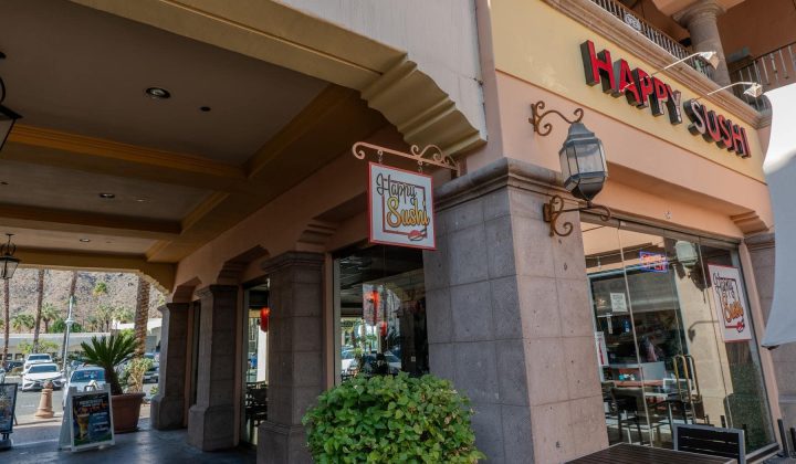 Entrance of "Happy Sushi" restaurant with a sign above the door, glass doors, and outdoor seating.