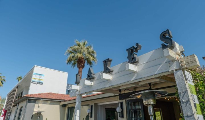 Outdoor view of a building with a white sign reading "JAKE'S" under a clear blue sky, flanked by a palm tree.