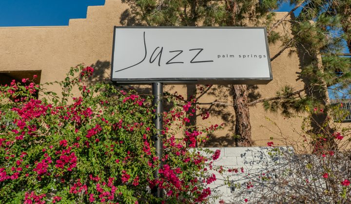 Sign reading "Jazz Palm Springs" stands amidst pink flowering plants and a tree, against a beige building backdrop.
