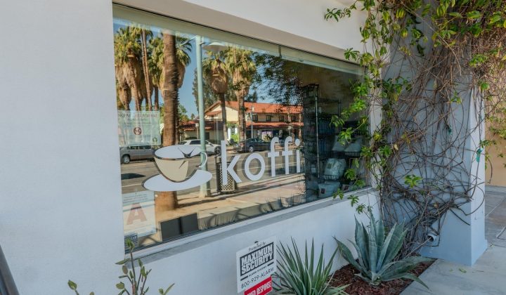 Cafe window with a cup logo and the word "koffi." Reflections of trees and a building visible. Plants and vines grow near the entrance. Nearby sign mentions Maximum Escapacity.