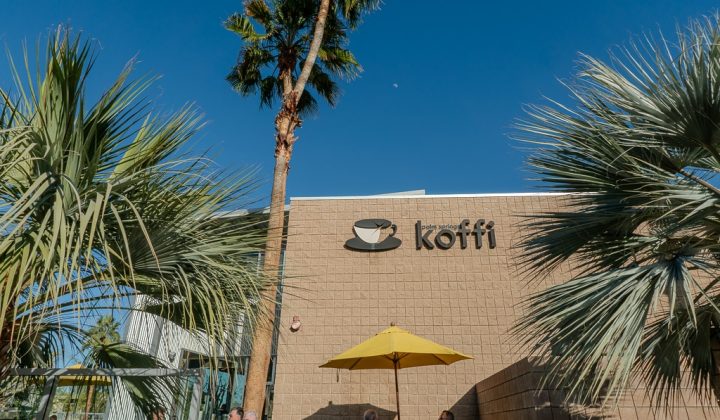Exterior of a coffee shop with the sign "koffi" against a blue sky. Palm trees flank the building, and people sit at a table under a yellow umbrella in the foreground.