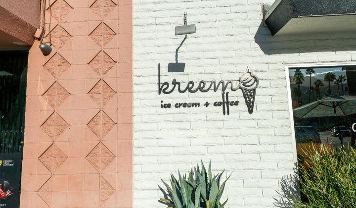 Exterior of a cafe with a sign reading "kreem ice cream + coffee" on a white brick wall. A small agave plant and turquoise chair are in the foreground.