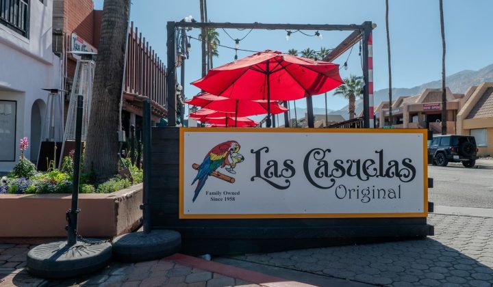 Outdoor sign for Las Casuelas Original restaurant, featuring a parrot illustration, with red umbrellas and palm trees in the background.