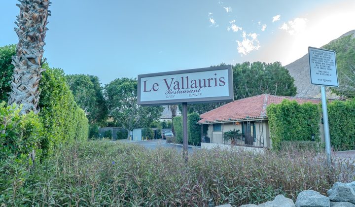 Sign for "Le Vallauris Restaurant" surrounded by greenery, with a red-roofed building in the background and a palm tree nearby.