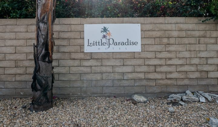 Sign on brick wall reads "Little Paradise Hotel" with palm tree logos; gravel and a tree in front.