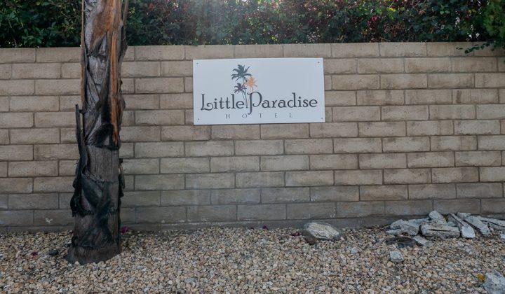 Sign on a brick wall reads "Little Paradise Hotel" above gravel and next to a tree, with foliage in the background.