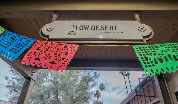 Sign reading "Low Desert Modern Mexican" with colorful papel picado banners under a wooden roof. Trees and sky are visible in the background.