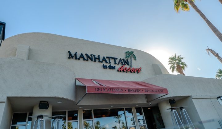 Front view of Manhattan in the Desert restaurant with palm trees around, featuring a beige facade and a red awning with signage for delicatessen, bakery, and restaurant.