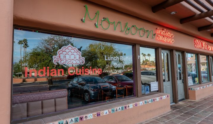 Exterior of Monsoon Indian Cuisine restaurant with large windows displaying signs for "Indian Cuisine" and "Lunch Buffet Daily." Reflections of parked cars and trees are visible on the glass.