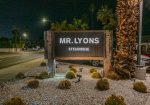 Sign for "Mr. Lyons Steakhouse" at night, surrounded by small cactus plants and a well-lit street in the background.
