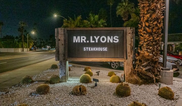 Sign for "Mr. Lyons Steakhouse" at night, surrounded by small cactus plants and a well-lit street in the background.