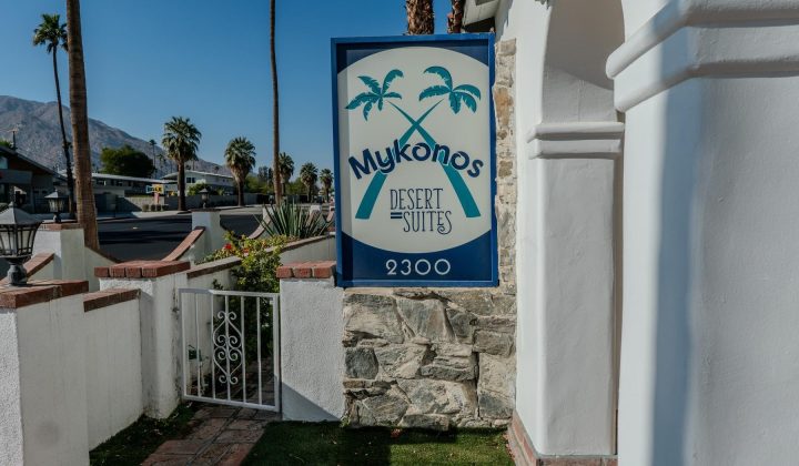 Sign reading "Mykonos Desert Suites" with crossed palm trees, attached to a white stone building, near a gate. Background shows palm trees and mountains under a clear blue sky.