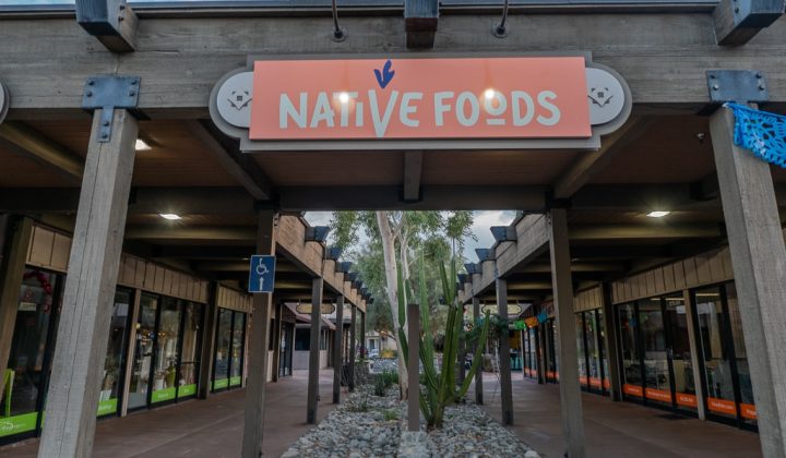 Exterior view of a restaurant with a sign reading "Native Foods." Pathway lined with pebbles and plants, flanked by shops under a wooden pergola.
