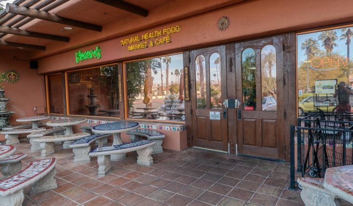 Exterior of a natural health food market and cafe, featuring tile benches, a large wooden door, and a neon "Open" sign.