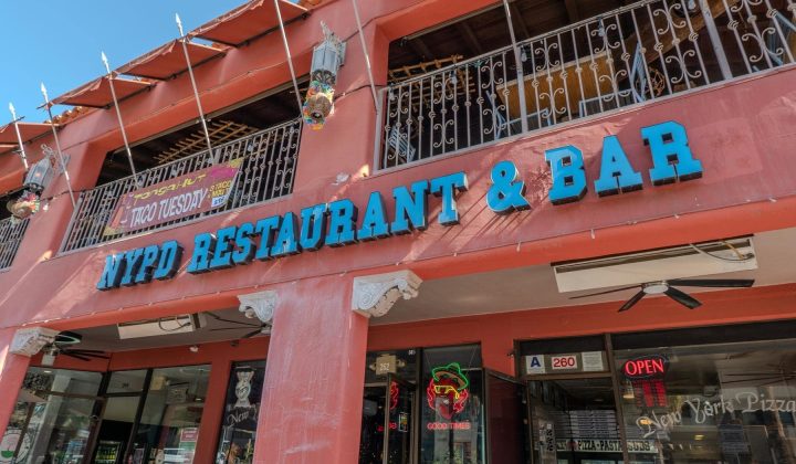Red building exterior with a sign reading "NYPD Restaurant & Bar" in blue letters. Patio area above with hanging lights. Neon "Open" sign and "Good Times" sign below.
