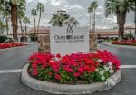 Entrance sign of Omni Rancho Las Palmas Resort, surrounded by red and pink flowers, with palm trees and a cloudy sky in the background.